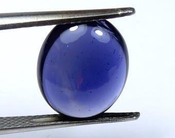 May include: A deep blue, oval-shaped gemstone is held by a pair of metal tweezers. The gem has a smooth, polished surface with a gradient of color, transitioning from a darker shade at the edges to a lighter hue in the center. The gemstone is approximately 1 inch in length.