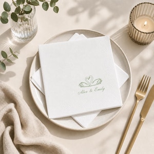 May include: A square white paper napkin featuring a green line-art illustration of two swans forming a heart shape above the printed text Alex & Emily in a cursive font, centered on a round ceramic plate with a smooth, light-colored finish.