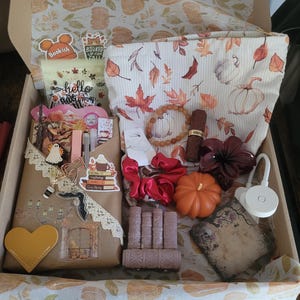 Könnte beinhalten: Eine Geschenkbox mit einem Beutel mit Kürbismuster, einem roten Haargummi, einer kürbisförmigen Kerze, einer buchförmigen Kerze, einem Perlenarmband, einer dunkelroten Blumenverzierung, einem herzförmigen Anhänger und Aufklebern.