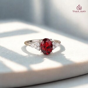 May include: A silver ring with a large, oval-cut, deep red gemstone as the focal point, complemented by smaller, clear stones. The ring has a slender band and is set against a soft, neutral backdrop. This is a piece of jewellery.