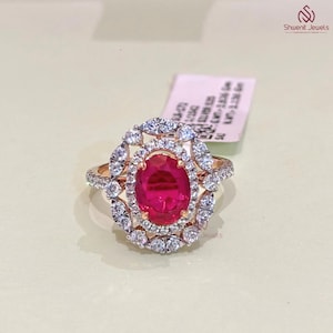 Puede incluir: Un anillo de oro rosa con una gran piedra central de rubí ovalada, rodeada por un halo de diamantes redondos más pequeños. Diamantes adicionales adornan la banda del anillo, creando un diseño brillante y elegante. El anillo está sobre un fondo neutro.