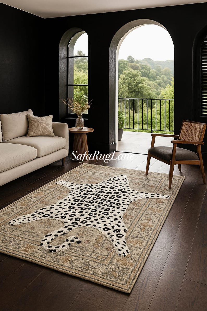 May include: A beige area rug with a leopard print rug on top. The leopard rug has black spots on a cream background. The area rug has a floral border. The room has a sofa, chair, and a view of trees.
