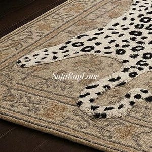 May include: A beige area rug with a leopard pattern in black and white. The rug has a detailed border and is placed on a dark wood floor. The SafaRugLane logo is visible on the rug.