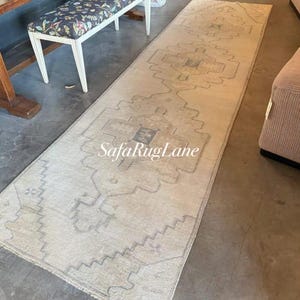 Könnte beinhalten: Ein langer, rechteckiger Teppich mit einem beige-grauen geometrischen Muster. Der Teppich hat ein zentrales Design mit sich wiederholenden Formen und einem Rand. Der Text "SafaRugLane" ist auf dem Teppich zu sehen.