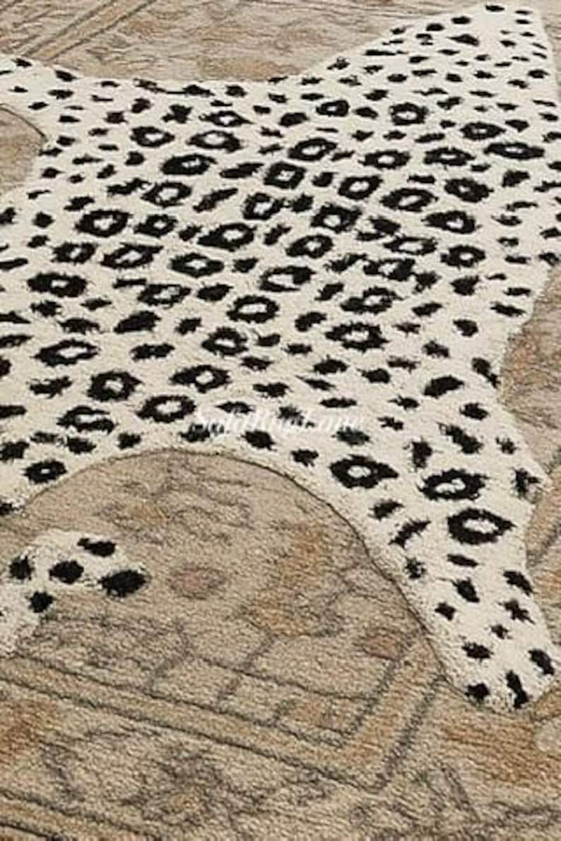 May include: A cream-colored rug with a black leopard print pattern. The rug is shaped like an animal hide and is placed on top of a patterned beige rug.