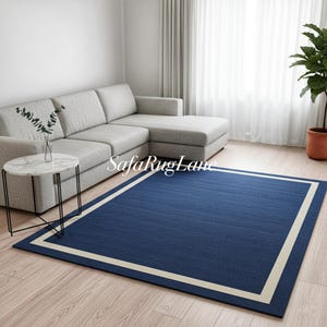 Könnte beinhalten: Ein rechteckiger marineblauer Teppich mit cremefarbenem Rand wird in einem Wohnzimmer gezeigt. Der Teppich liegt auf einem Holzboden, mit einem grauen Ecksofa und einem kleinen Beistelltisch im Hintergrund. Der Text "SafaRugLane" ist auf dem Teppich zu sehen.