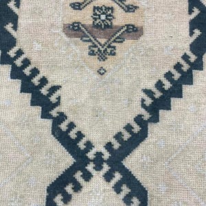 Könnte beinhalten: Ein gewebter Teppich mit beigem Hintergrund und einem markanten geometrischen Rautenmuster in Dunkelanthrazit. Die Mitte der Raute enthält ein detailliertes Motiv in Dunkelanthrazit und Braun. Der Text SafaRugstane ist mittig sichtbar.