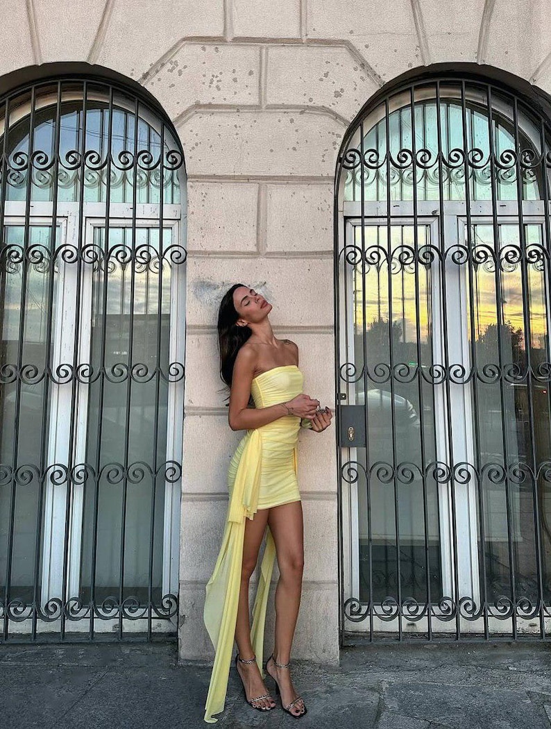 May include: A strapless, pale yellow mini dress with a flowing, asymmetrical train. The dress is paired with strappy high heels. The background features arched windows with decorative ironwork and a light-colored stone wall.
