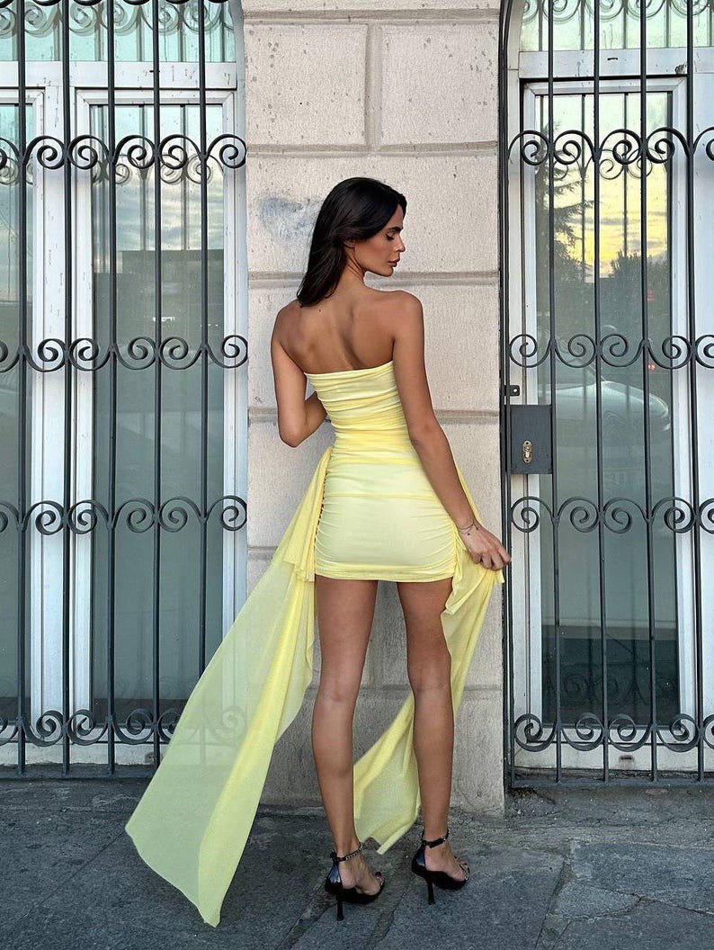 May include: A strapless, pale yellow dress with a draped, flowing train. The short, ruched dress is paired with black high-heeled sandals. The model is standing in front of a building with ornate ironwork.