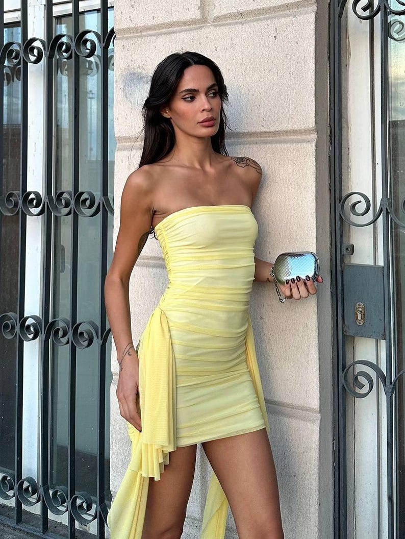 May include: A strapless, pale yellow mini dress with a draped, asymmetrical skirt. The dress is made of a sheer, textured fabric. A small, silver clutch is held in the hand.
