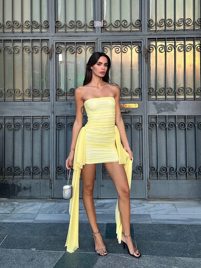 May include: A strapless, pale yellow dress with a ruched bodice and flowing side panels. The dress is paired with black high-heeled sandals and a small silver purse. The background features a dark gray metal door.