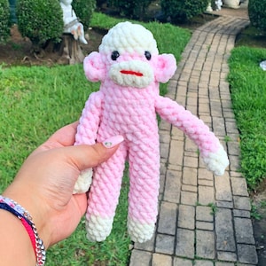 May include: A pink crocheted monkey toy with white yarn accents. The monkey has a white face with pink eyes and a red mouth.