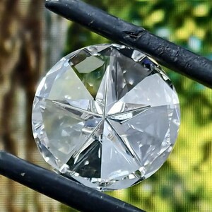 May include: A clear, round-cut diamond with a star-shaped facet pattern. The diamond is held by black tweezers against a blurred green and brown background. Jewelry and gemstone.