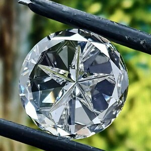 May include: A large, round, faceted diamond held by black tweezers. The diamond has a star-like pattern in the center and reflects light, creating a brilliant sparkle. The background is a blurred green.