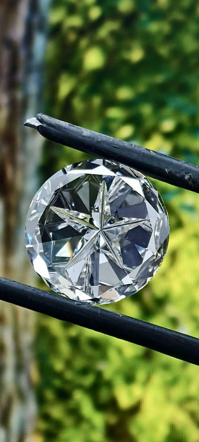 May include: A large, round, faceted diamond held by black tweezers. The diamond has a star-like pattern in the center and reflects light, creating a brilliant sparkle. The background is a blurred green.