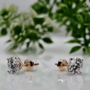 May include: A pair of diamond stud earrings. Each earring features a round-cut, clear diamond set in a rose gold setting. The earrings are displayed on a white surface with a blurred green and white background.