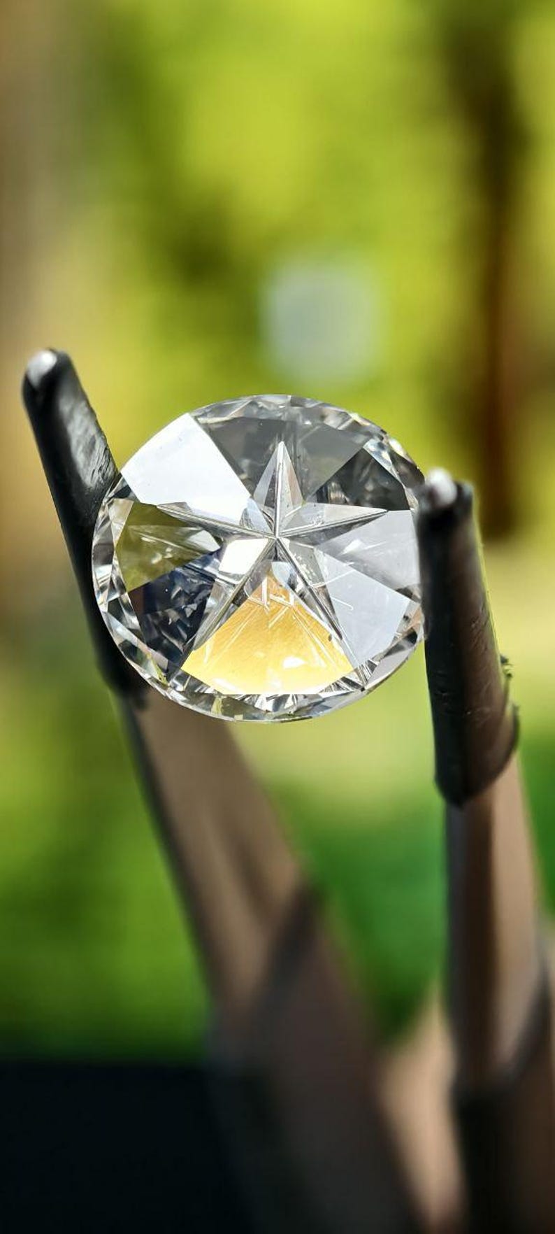 May include: A clear, faceted diamond held by black metal prongs. The diamond has a star-shaped design in the center and reflects light, creating a brilliant sparkle. The background is a blurred green.