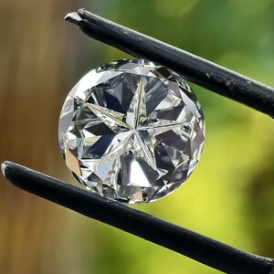 May include: A round, brilliant-cut diamond held by black tweezers. The diamond sparkles with multiple facets, reflecting light. The background is a soft, blurred green and brown.