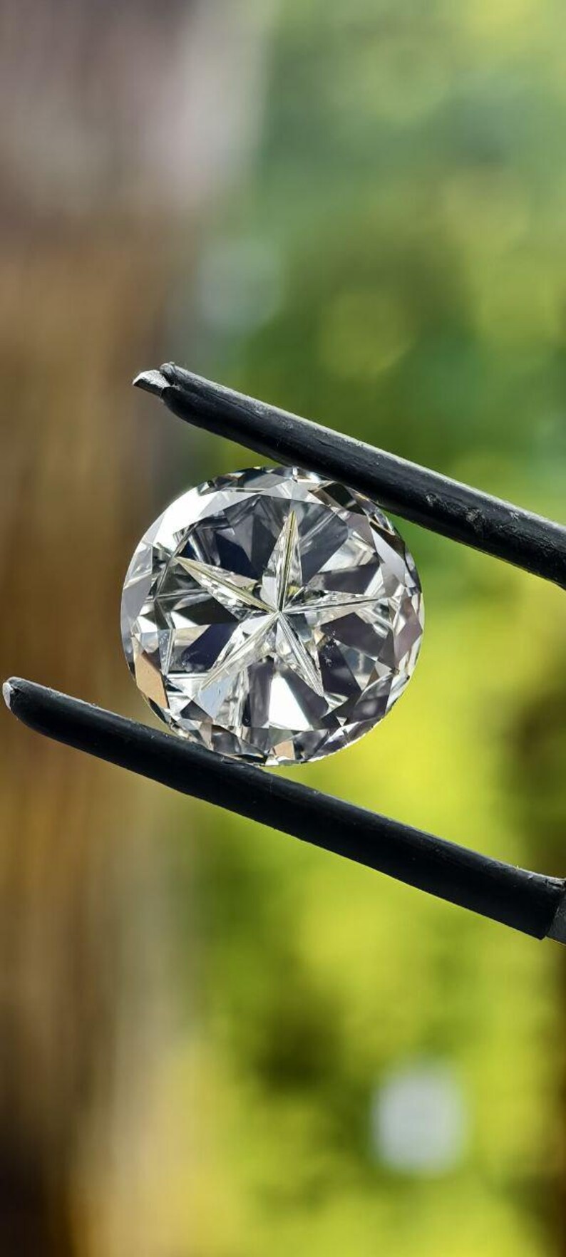 May include: A round, brilliant-cut diamond held by black tweezers. The diamond sparkles with multiple facets, reflecting light. The background is a soft, blurred green and brown.