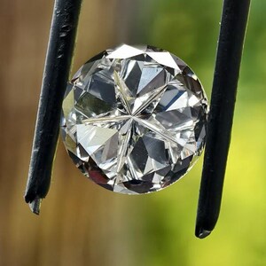 May include: A round, faceted diamond held by black-tipped tweezers. The diamond sparkles with numerous facets, reflecting light. The background is a blurred mix of green and brown.