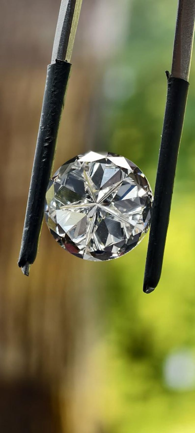 May include: A round, faceted diamond held by black-tipped tweezers. The diamond sparkles with numerous facets, reflecting light. The background is a blurred mix of green and brown.