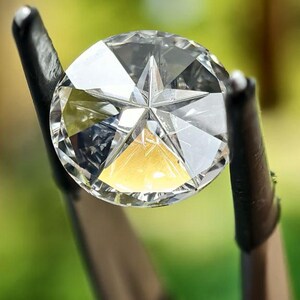 May include: A clear, faceted diamond held by black metal prongs. The diamond has a star-shaped design in the center. The background is a blurred green.