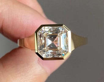 Puede incluir: Un anillo de oro con una gran piedra preciosa transparente de talla cuadrada. La piedra preciosa está engastada en un bisel de oro, y la banda es de oro pulido simple. El anillo se sostiene sobre un fondo neutro.