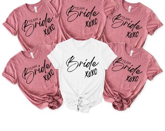 May include: A collection of six t-shirts, featuring a white tee and five pink tees. The white shirt has the word "Bride" in a cursive font, with "xoxo" below. The pink shirts say "Team Bride xoxo".