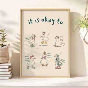 Könnte beinhalten: Ein gerahmter Kunstdruck mit cremefarbenem Hintergrund, der den Text 'it is okay to' über sechs Illustrationen von weißen Enten zeigt, die verschiedene Accessoires wie Schleifen und Stiefel tragen, jeweils mit kurzen ermutigenden Sätzen in Dunkelgrün.