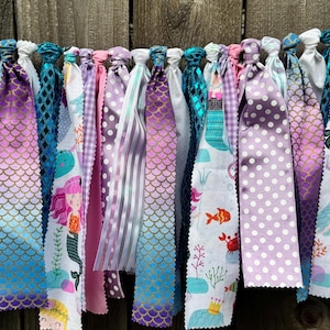 May include: A fabric rag garland featuring a variety of strips in shades of purple, teal, and pink. The fabric patterns include white polka dots, purple gingham, metallic scale prints, and illustrations of mermaids, fish, and sea life.