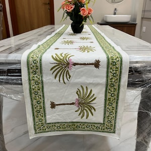 Könnte beinhalten: Weißer Tischläufer mit grünem und floralem Rand und Palmenmuster. Die Palmen haben grüne Wedel und rosa Blüten. Der Läufer liegt auf einem Marmortisch mit einer Blumenvase im Hintergrund.