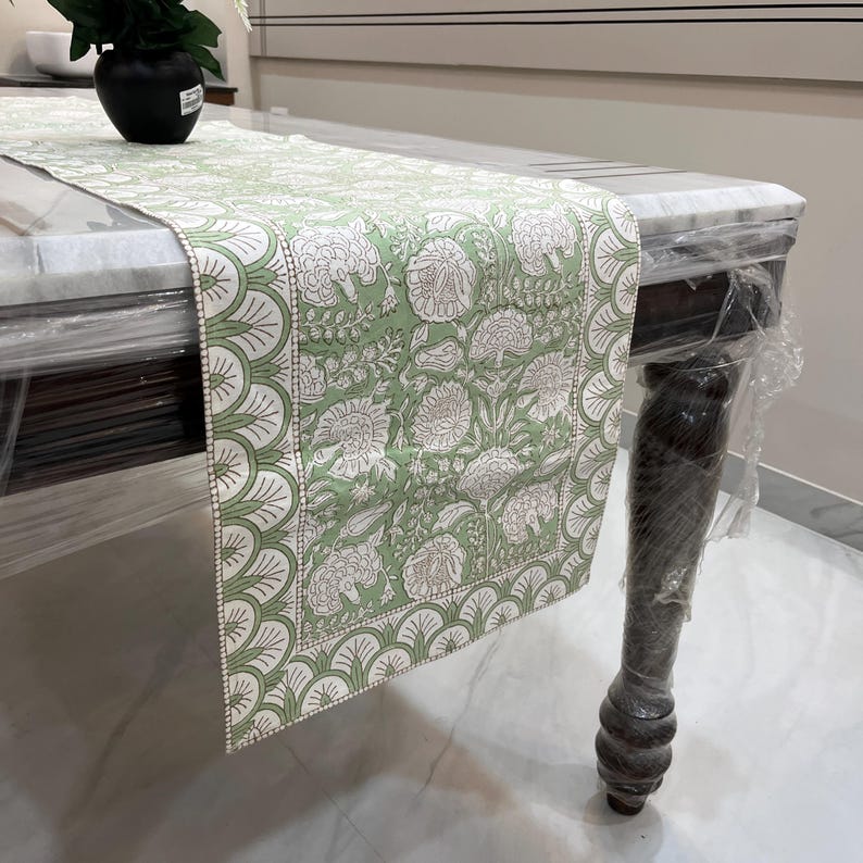 May include: A light green and white floral patterned table runner. The runner is draped over a marble-topped table with dark wooden legs. The floral design features detailed botanical elements.