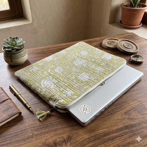 May include: A quilted laptop sleeve in a light olive green color featuring a white floral and botanical print. The sleeve has a zipper closure with a small matching tassel pull and contains a silver laptop with a circular sticker on the lid.