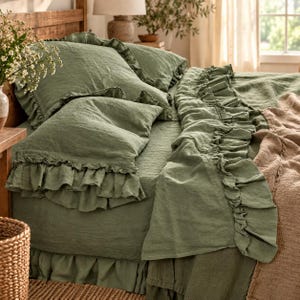 May include: A sage green bedding set featuring two ruffled pillowcases and a matching ruffled duvet cover. The fabric has a visible textured weave. A tan textured throw blanket is draped over the side of the bed, partially covering the green bedding.