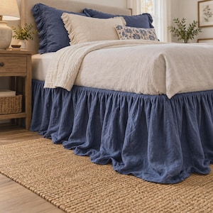May include: A blue linen bed skirt with a gathered, ruffled design that drapes around the base of the bed. The bed is dressed with a cream-colored textured coverlet, two blue ruffled pillow shams, a cream pillow, and a blue patterned pillow.