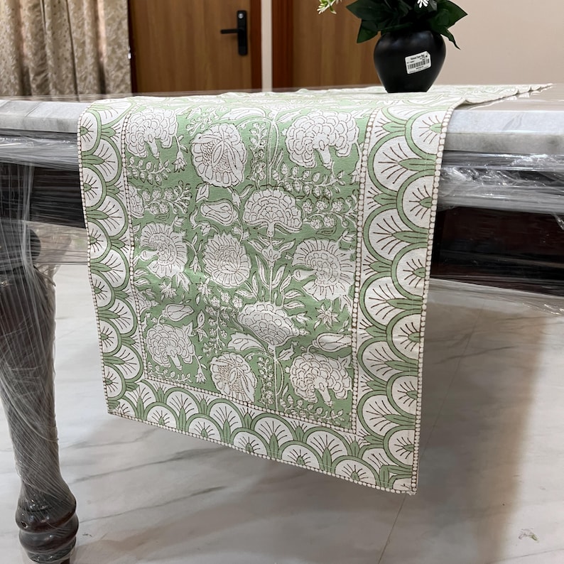 May include: A green and white floral patterned table runner. The runner features a repeating pattern of large white flowers and leaves on a light green background. The border has a scalloped design. The runner is draped over a table.