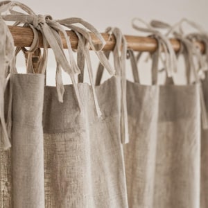 May include: A set of light beige linen curtains hanging from a wooden rod. The curtains feature fabric ties at the top that are knotted around the rod to secure the panels in place. The fabric has a visible woven texture and a natural, neutral colour.