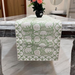 Pode incluir: Um caminho de mesa floral verde e branco. O corredor apresenta um padrão repetido de grandes flores e folhas brancas sobre um fundo verde claro. As bordas são aparadas com uma borda ondulada em branco e verde. Um vaso preto com flores está em cima.