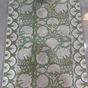May include: A rectangular green and white floral table runner. The runner features a repeating pattern of large white flowers and leaves on a green background. The edges are bordered with a scalloped design in white and green.