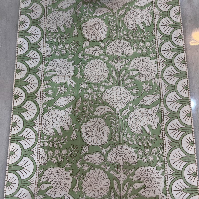 May include: A rectangular green and white floral table runner. The runner features a repeating pattern of large white flowers and leaves on a green background. The edges are bordered with a scalloped design in white and green.