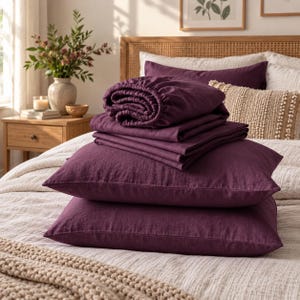 May include: A set of deep plum-colored bedding including two rectangular pillowcases and a folded fitted sheet. The fabric has a visible woven texture. The items are stacked neatly on top of a light-colored textured bedspread in a bedroom setting.