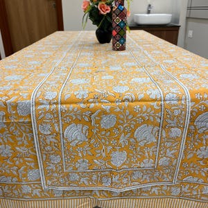 Puede incluir: Un mantel amarillo con un estampado floral en blanco y gris. El mantel tiene un borde con un diseño de rayas blancas. La mesa está decorada con un jarrón de flores y una botella decorativa.