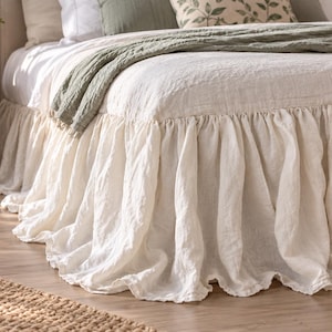 May include: A cream-coloured bed skirt featuring a long, gathered ruffle design that extends to the floor. The fabric has a visible, textured weave, creating a soft, draped appearance around the base of the bed frame, providing full coverage.