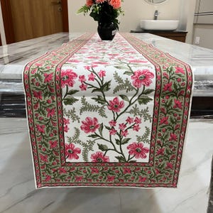 Può includere: Runner da tavolo bianco con un motivo floreale di fiori rosa e foglie verdi. Il runner ha un bordo rosa e verde. Un vaso nero con fiori è posto sul runner da tavolo.