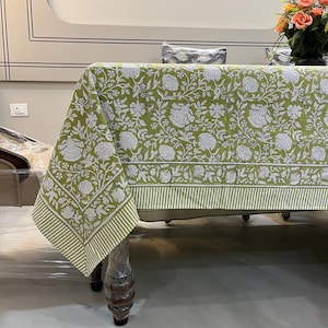 Puede incluir: Un mantel verde floral con patrones de flores y hojas blancas. El mantel tiene un borde a rayas en verde y blanco. La mesa es de madera y tiene patas decorativas.