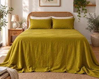 May include: A bed featuring a solid olive green bedspread and two matching rectangular pillows. The bed has a curved headboard made of light brown wood and woven cane, positioned against a white wall with a framed botanical print above it.