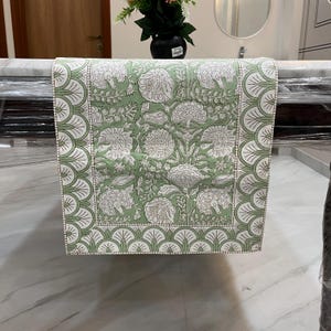 May include: A green and white floral patterned table runner. The runner features a repeating floral design in white on a green background. The border has a scalloped design in white and green. This decorative table runner is suitable for dining tables.