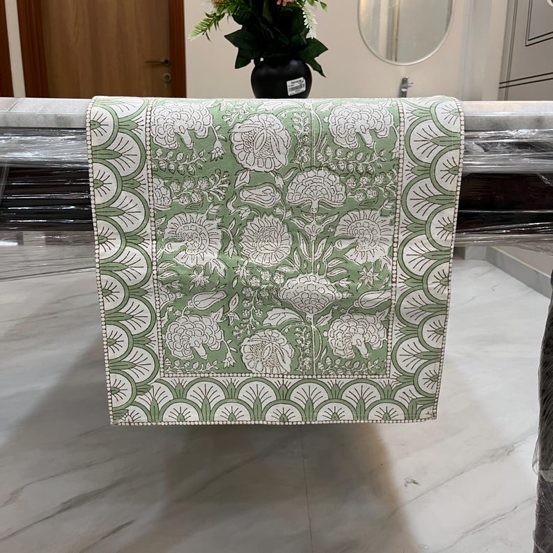May include: A green and white floral patterned table runner. The runner features a repeating floral design in white on a green background. The border has a scalloped design in white and green. This decorative table runner is suitable for dining tables.