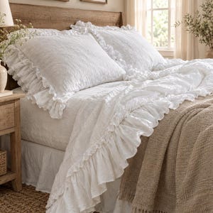 Puede incluir: Un juego de cama blanco que incluye una funda nórdica y fundas de almohada a juego con bordes de volantes. La tela tiene una apariencia texturizada y arrugada. Una manta tejida de color tostado cubre la esquina de la cama.