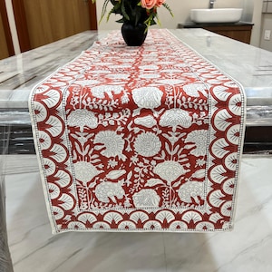 May include: A red and white floral table runner. The runner features a repeating pattern of large white flowers and foliage on a red background. The border has a scalloped design. The runner is placed on a marble table.